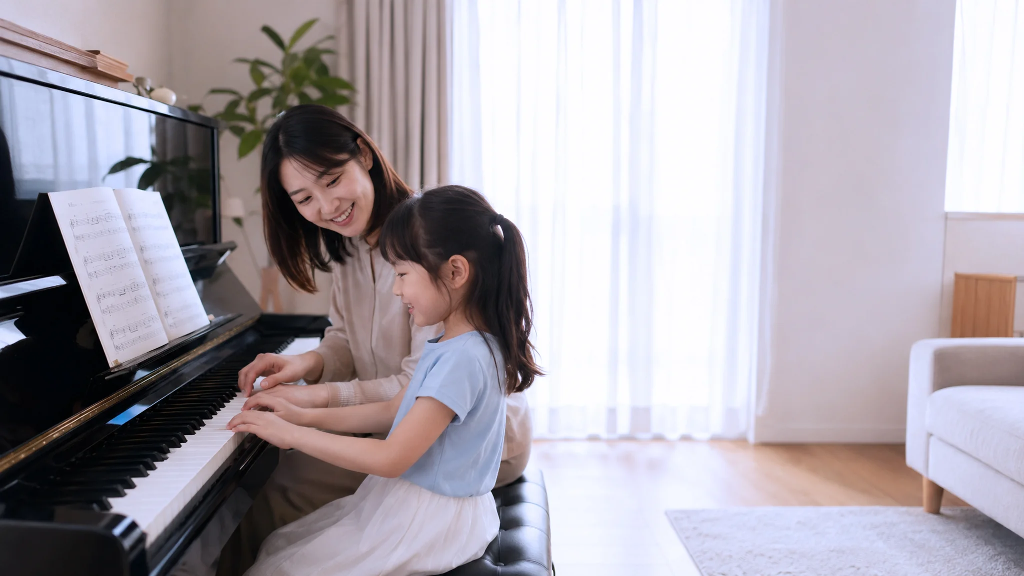 A piano teacher and a young girl smiling together at an upright piano in a bright, modern home